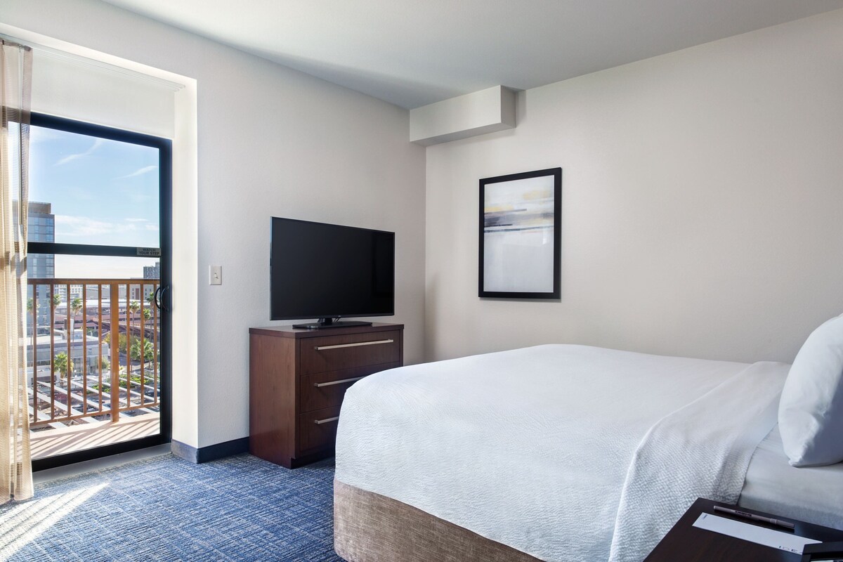 A hotel room with a bed, a television on a wooden dresser, a framed picture on the wall, and a balcony with a city view.