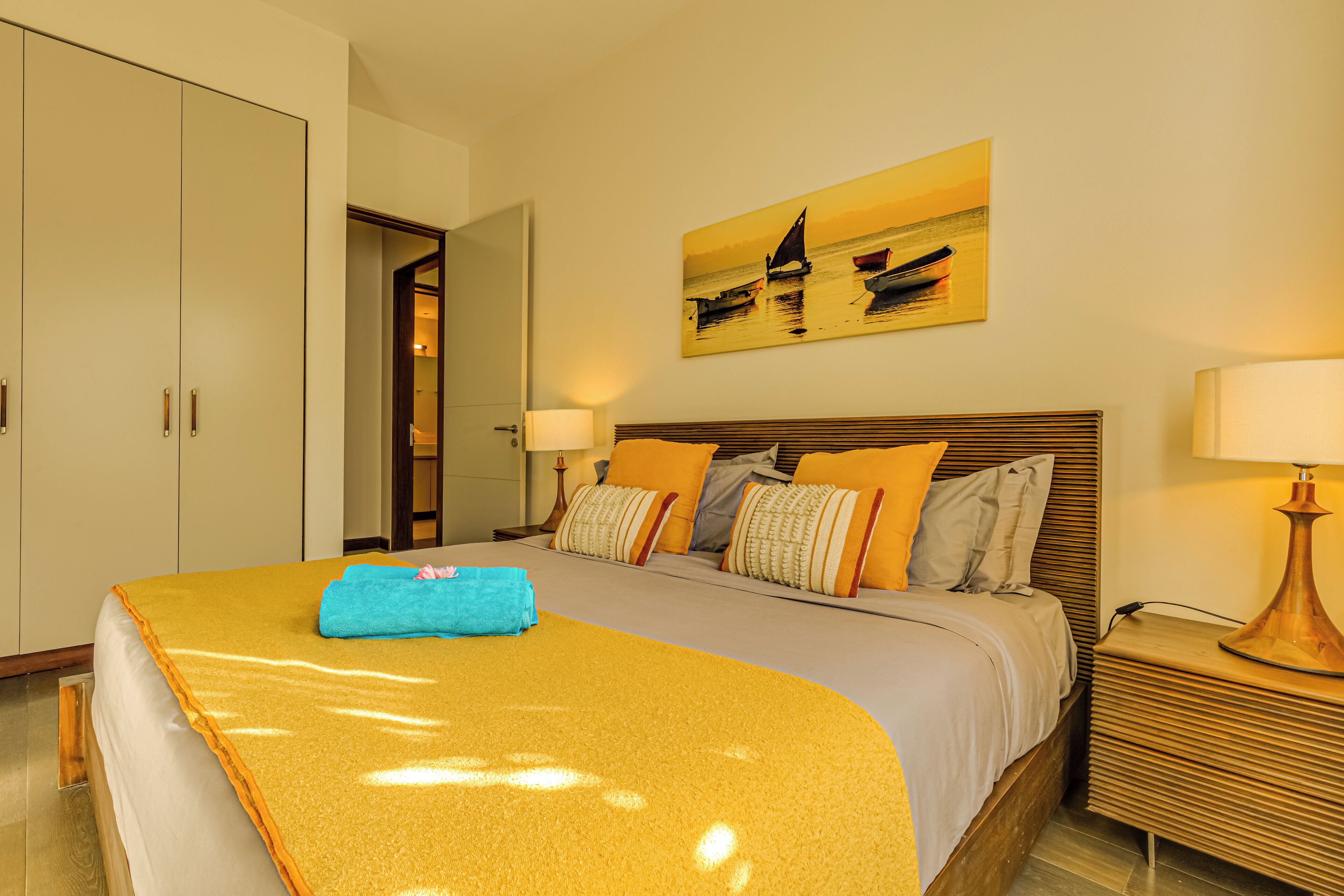 A hotel room with a bed, a painting of boats on the wall, a nightstand, and a door leading to another room.