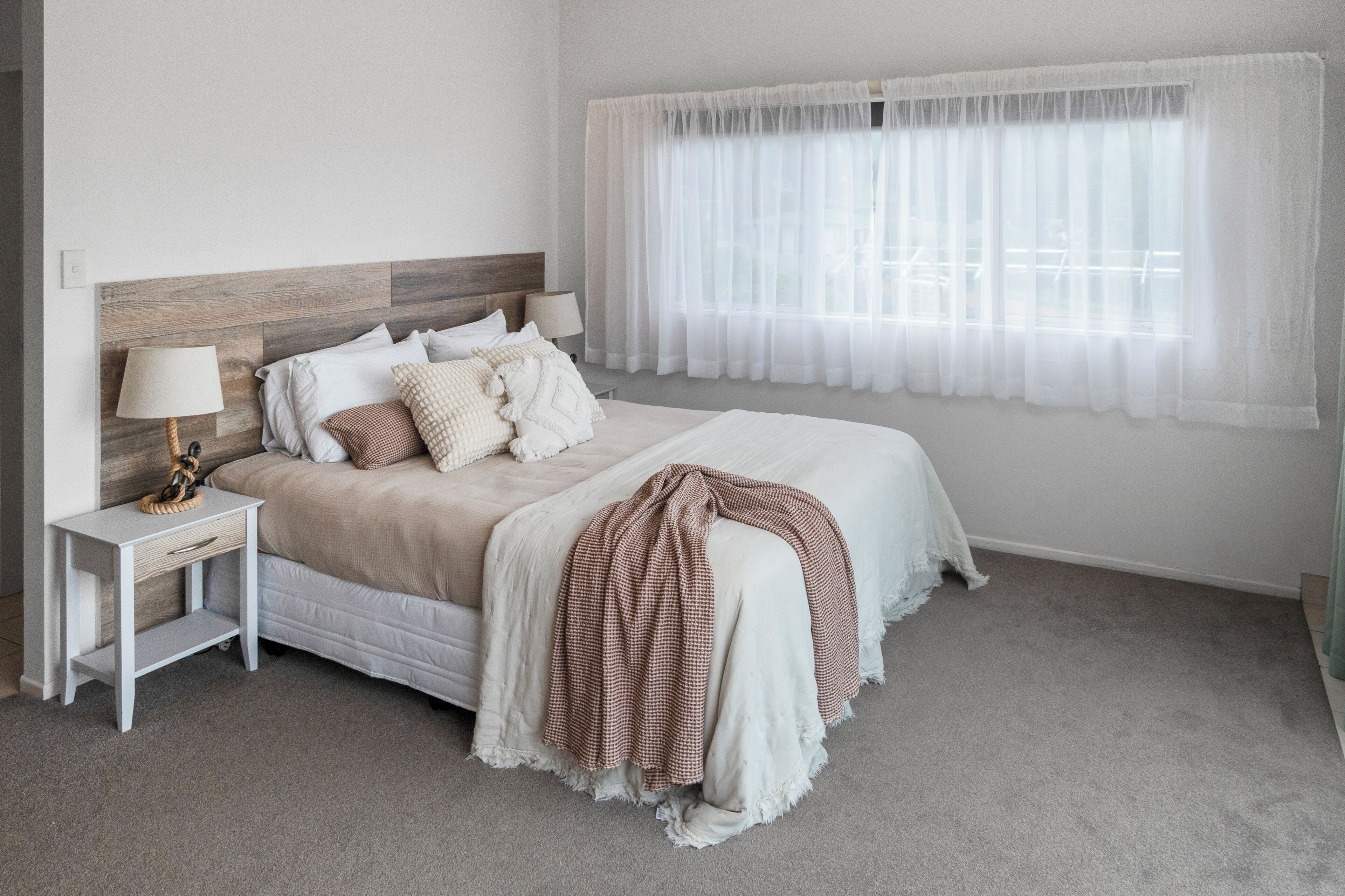 A bedroom with a bed, nightstand, lamp, and window with sheer curtains.