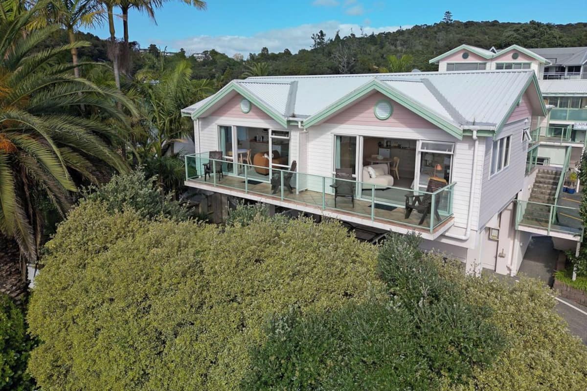 A two-story house with a balcony, surrounded by lush greenery and palm trees.