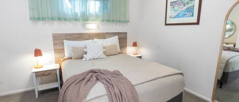 A neatly made bed with a checkered throw, two bedside lamps, a nightstand, a framed picture on the wall, and a large mirror.