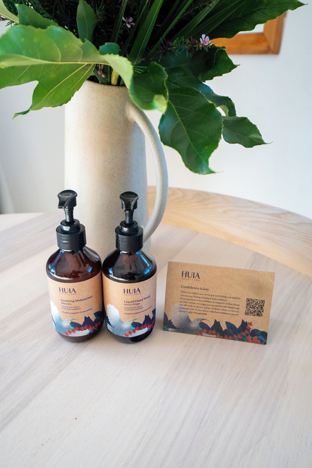 Dos botellas de jabón de manos HUIA y una tarjeta sobre una superficie de madera, con una planta al fondo.