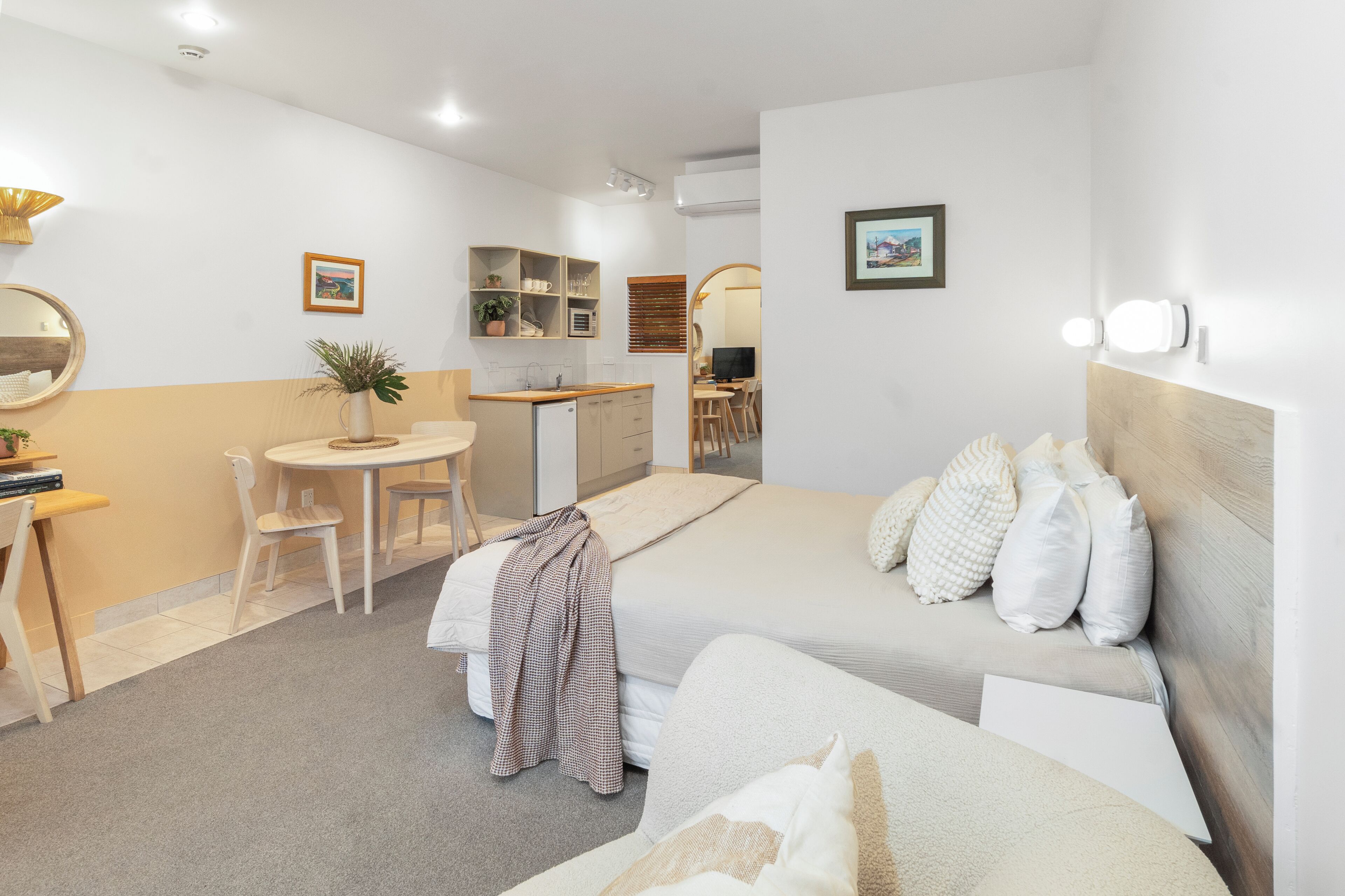 Une chambre d’hôtel moderne équipée d’un lit, d’une table de chevet et d’un petit bureau avec une chaise. Elle comprend également un coin cuisine avec un micro-ondes et un évier.