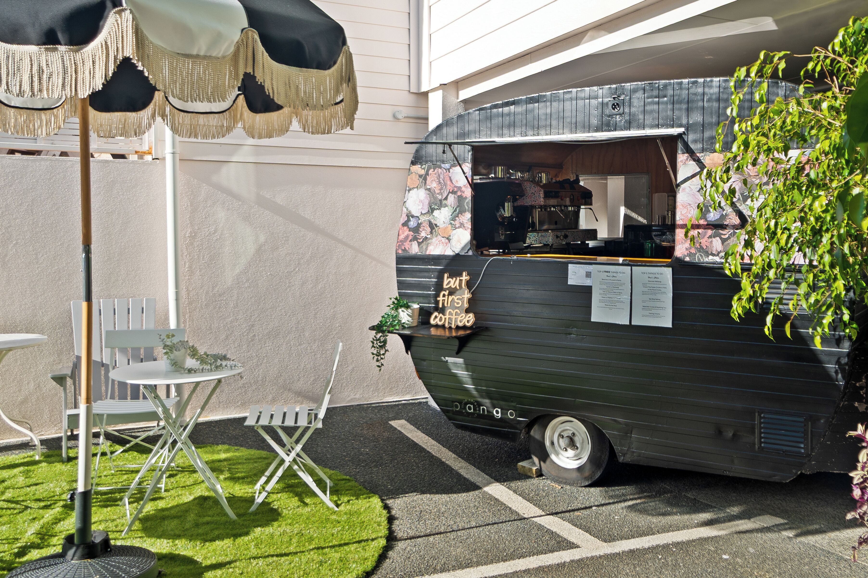 A vintage vehicle with a sign reading "but first coffee" parked in a courtyard with a table and chairs.
