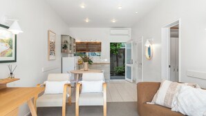 A modern living room with a brown sofa, a wooden coffee table, and a white armchair. There is a kitchen in the background with a window and a plant. - ASURE Admirals View Motel (Paihia)