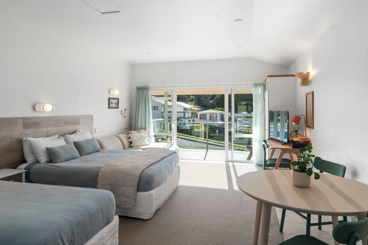 A modern bedroom with two beds, a dining table, and a view of a residential area.