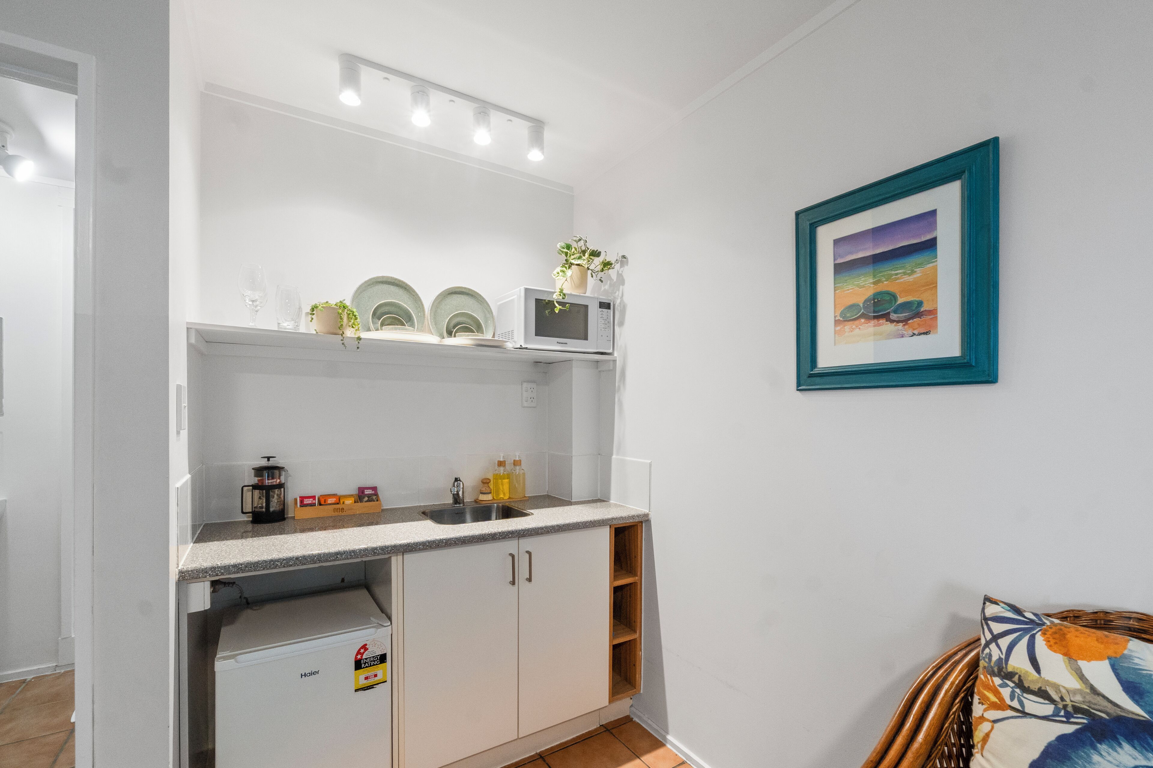 A compact kitchen with white cabinets, a microwave, and a sink. There is a framed picture on the wall and a wicker chair in the corner.