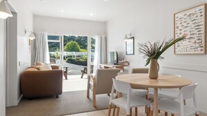 A modern living room with a sofa, a dining table with chairs, a TV, and a large window with a view of greenery. - ASURE Admirals View Motel (Paihia)
