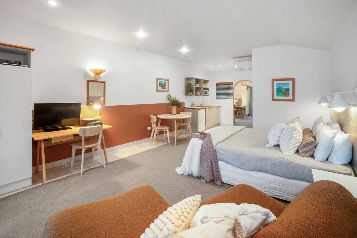 A modern hotel room with a bed, a desk with a computer, a TV, a dining area, and a kitchenette.