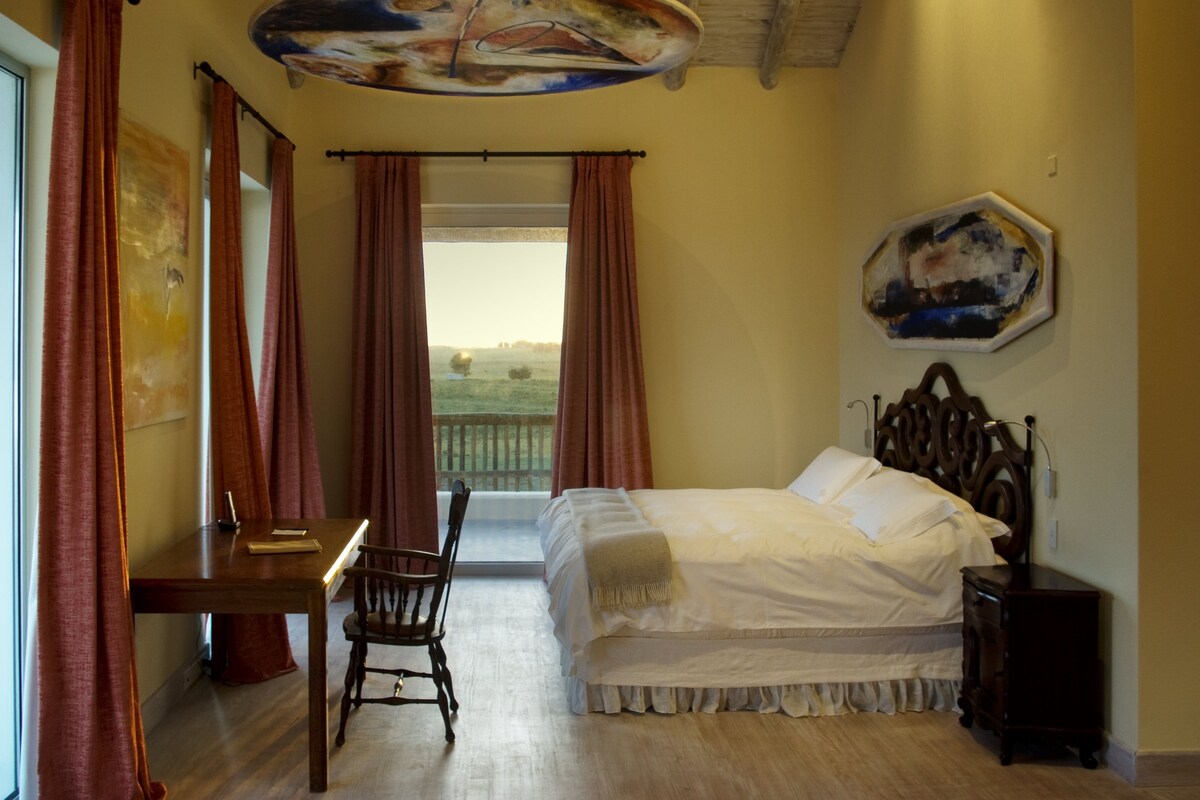 A bedroom with a wooden bed, a desk, and a chair. There is a window with curtains, a painting on the wall, and a view of the outdoors.
