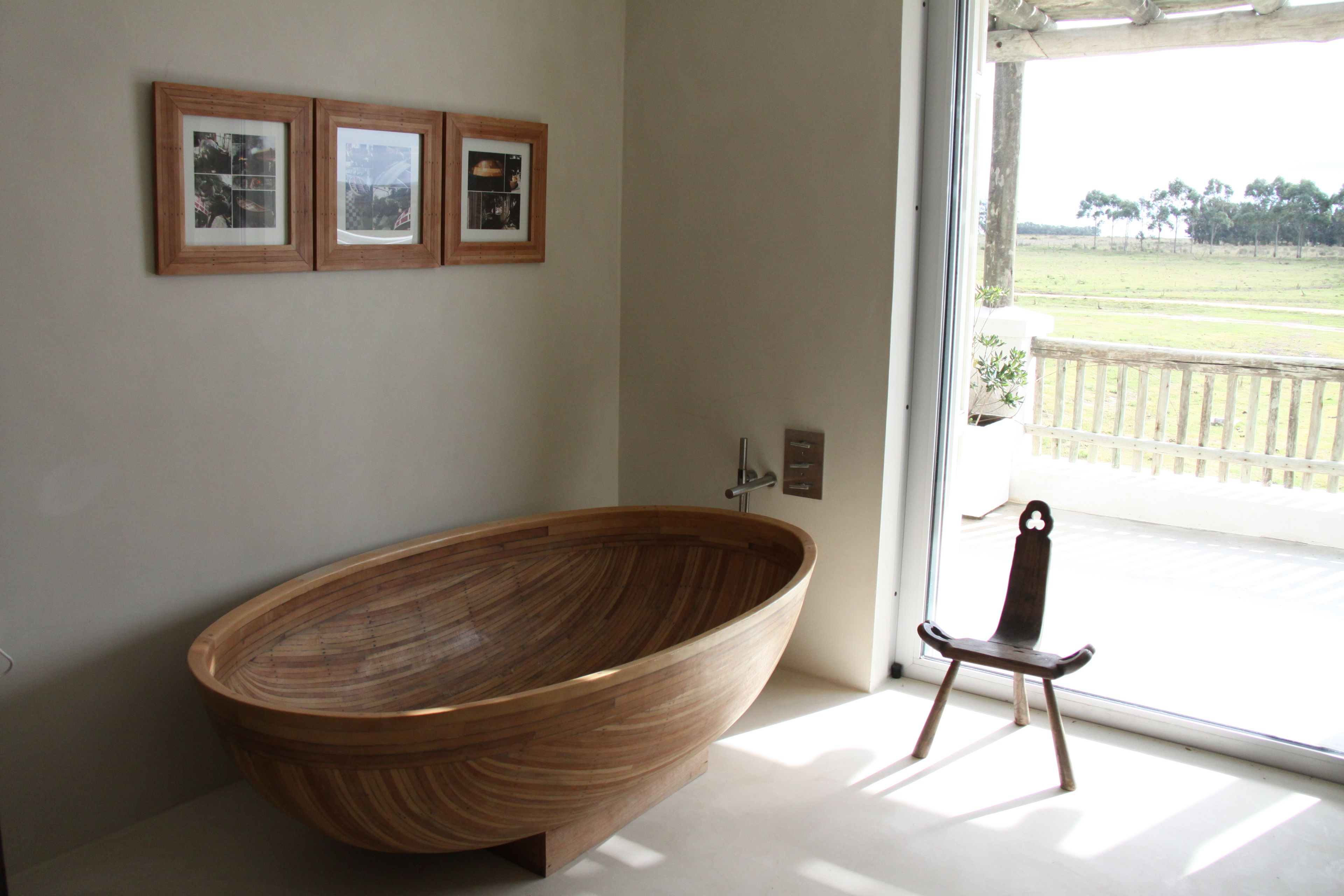 Un baño moderno con una bañera de madera grande, cuadros en la pared y vistas a un paisaje verde a través de un ventanal amplio.