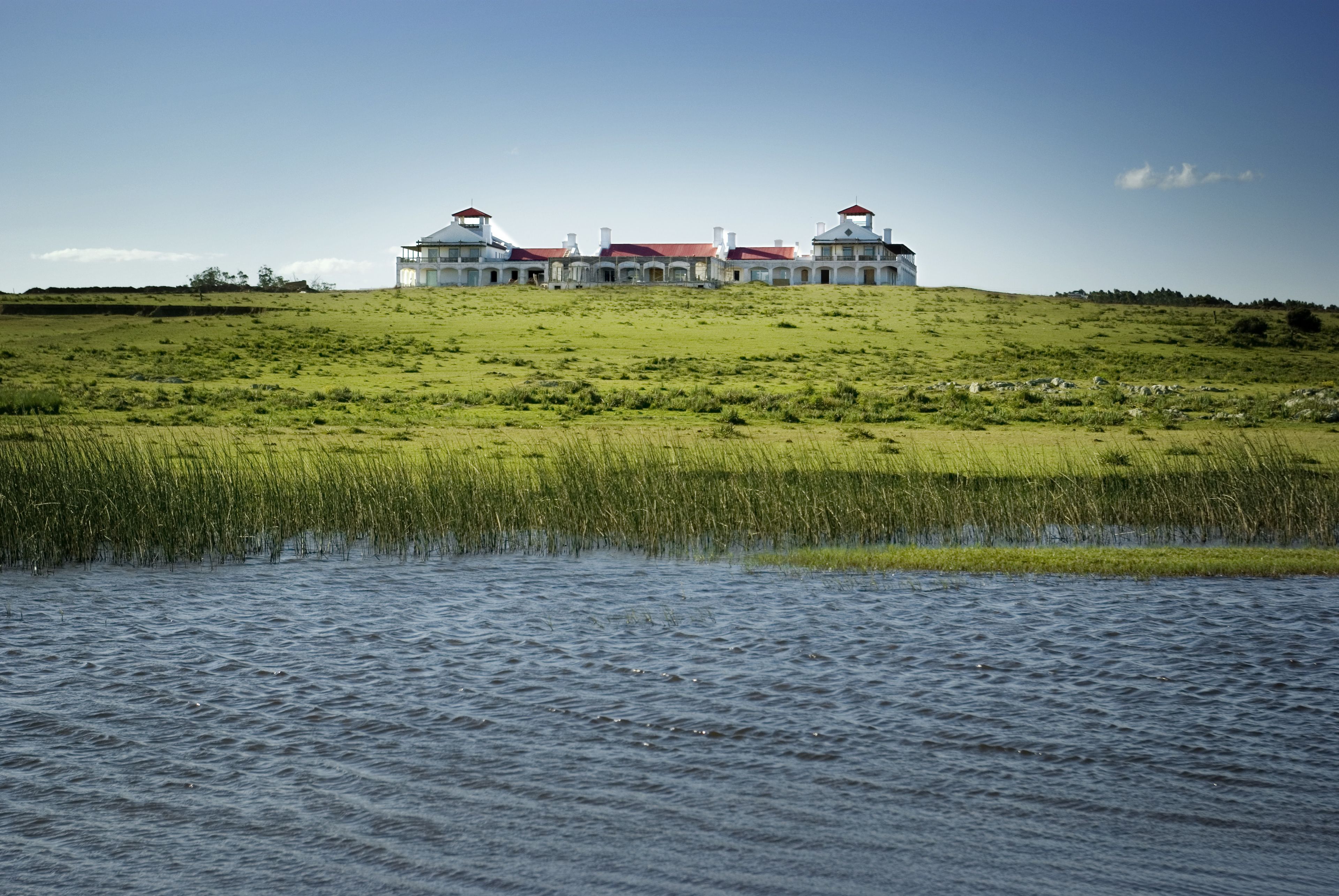 A large, white building with red roofs on a grassy hill, overlooking a body of water.