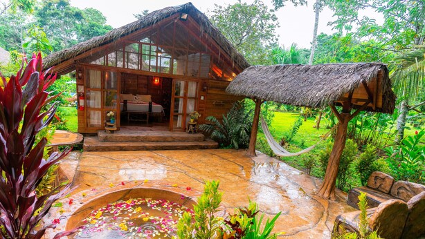 A rustic wooden house with a thatched roof, a stone-paved courtyard, and a small pond with floating flowers. - Suchipakari Jungle Lodge (Misahualli)