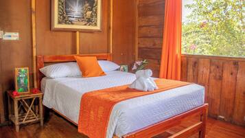 A wooden-framed bed with white bedding and an orange throw blanket, a small bedside table with a plant, and a framed picture on the wall.