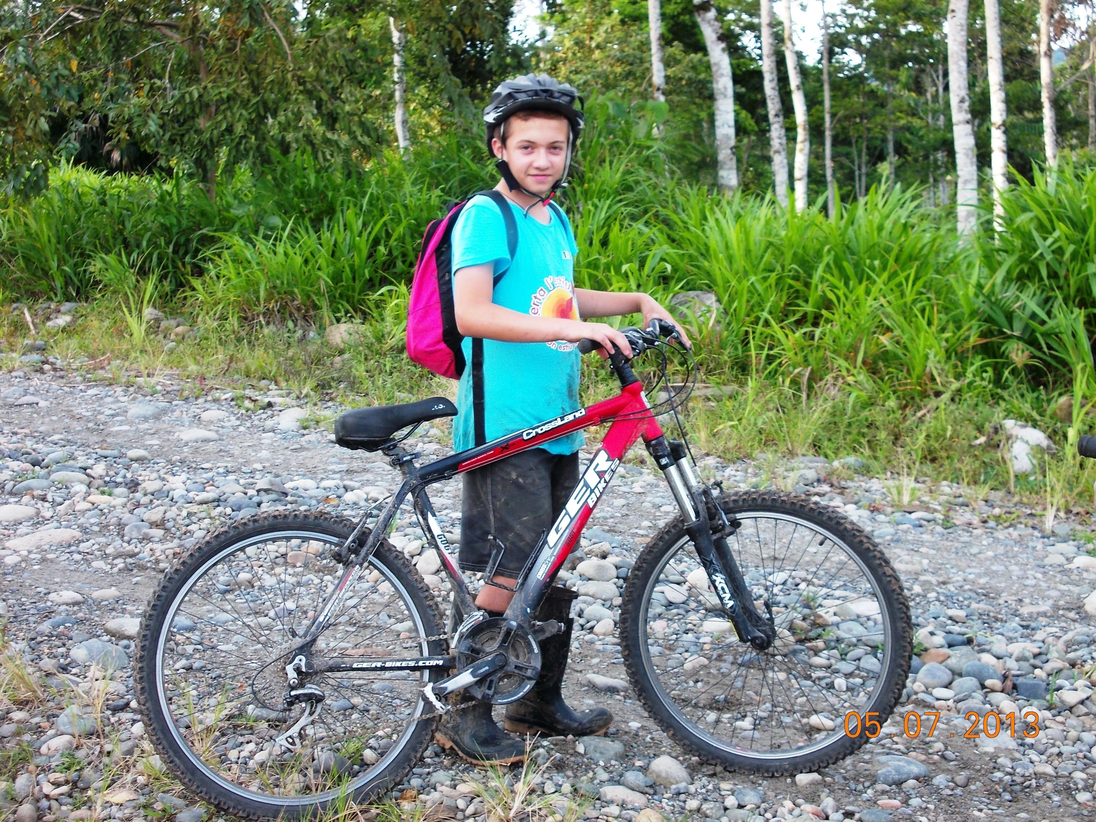 Uma pessoa ao lado de uma bicicleta de montanha numa trilha de cascalho, com vegetação ao fundo.