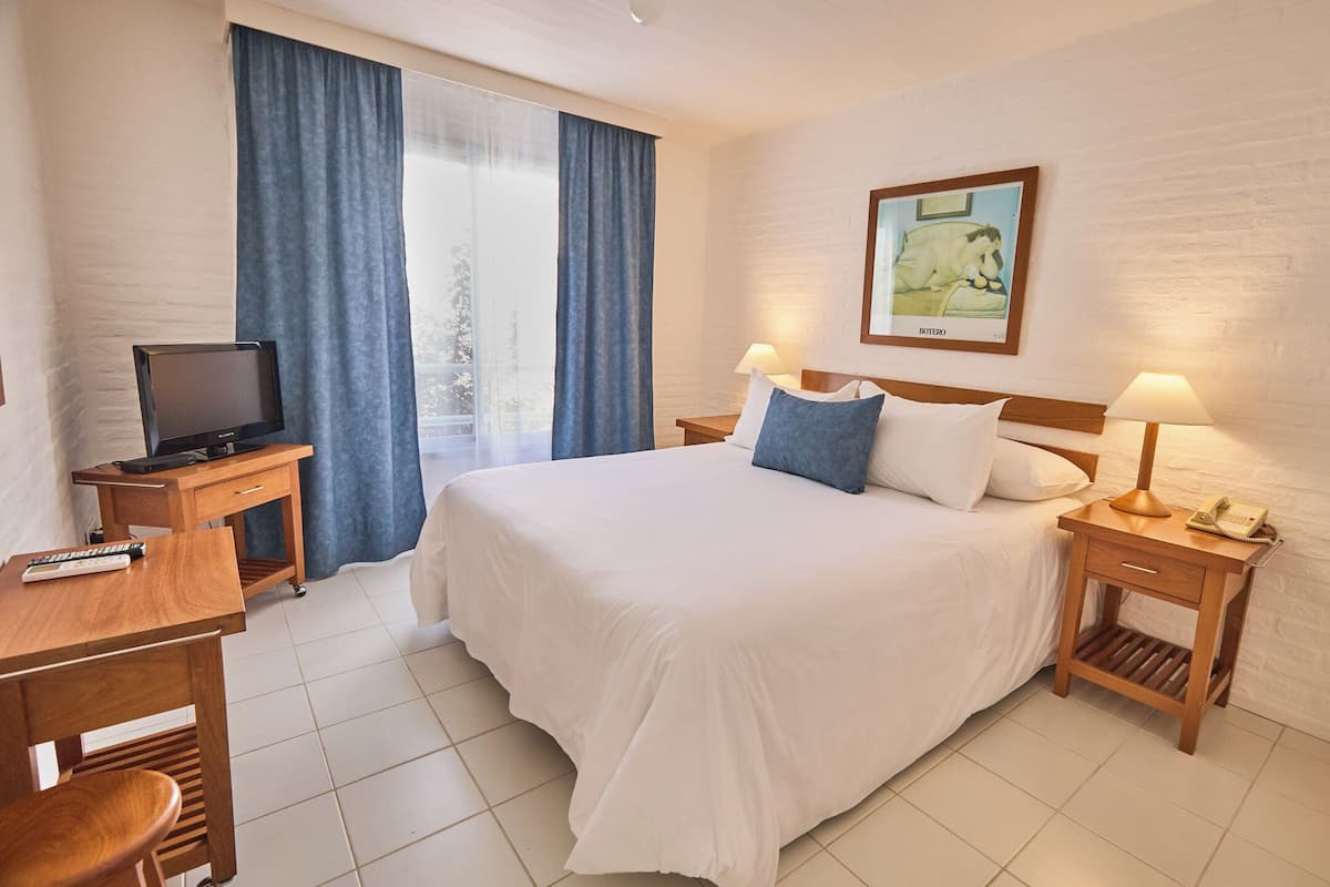 Habitación de hotel con cama, dos mesitas de noche con lámparas, escritorio con televisor y ventana con cortinas.