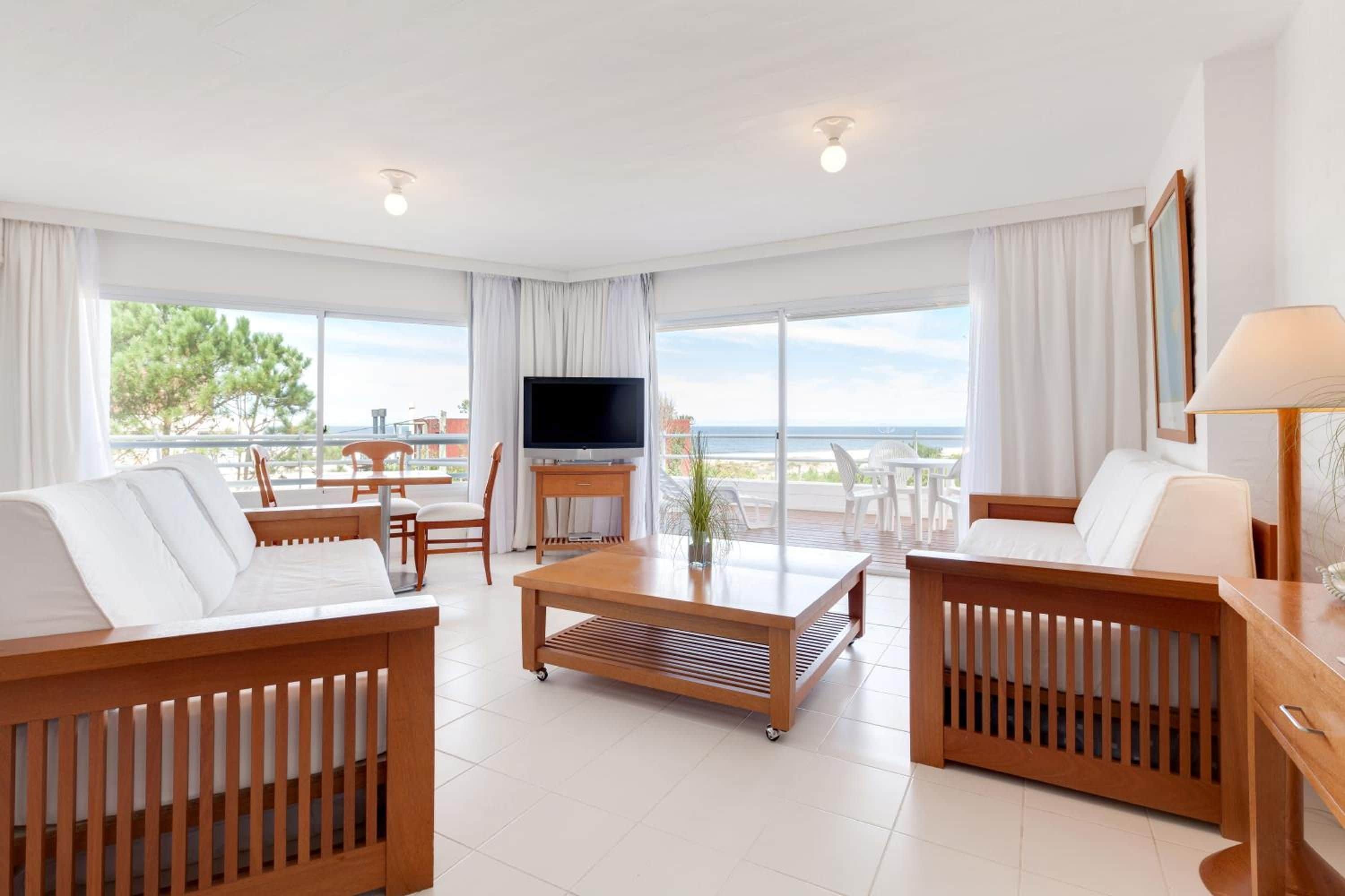 A modern hotel room with a balcony, a flat-screen TV, and a dining area with a table and chairs.