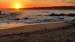Une plage au coucher du soleil, avec des vagues, des rochers et une côte lointaine.