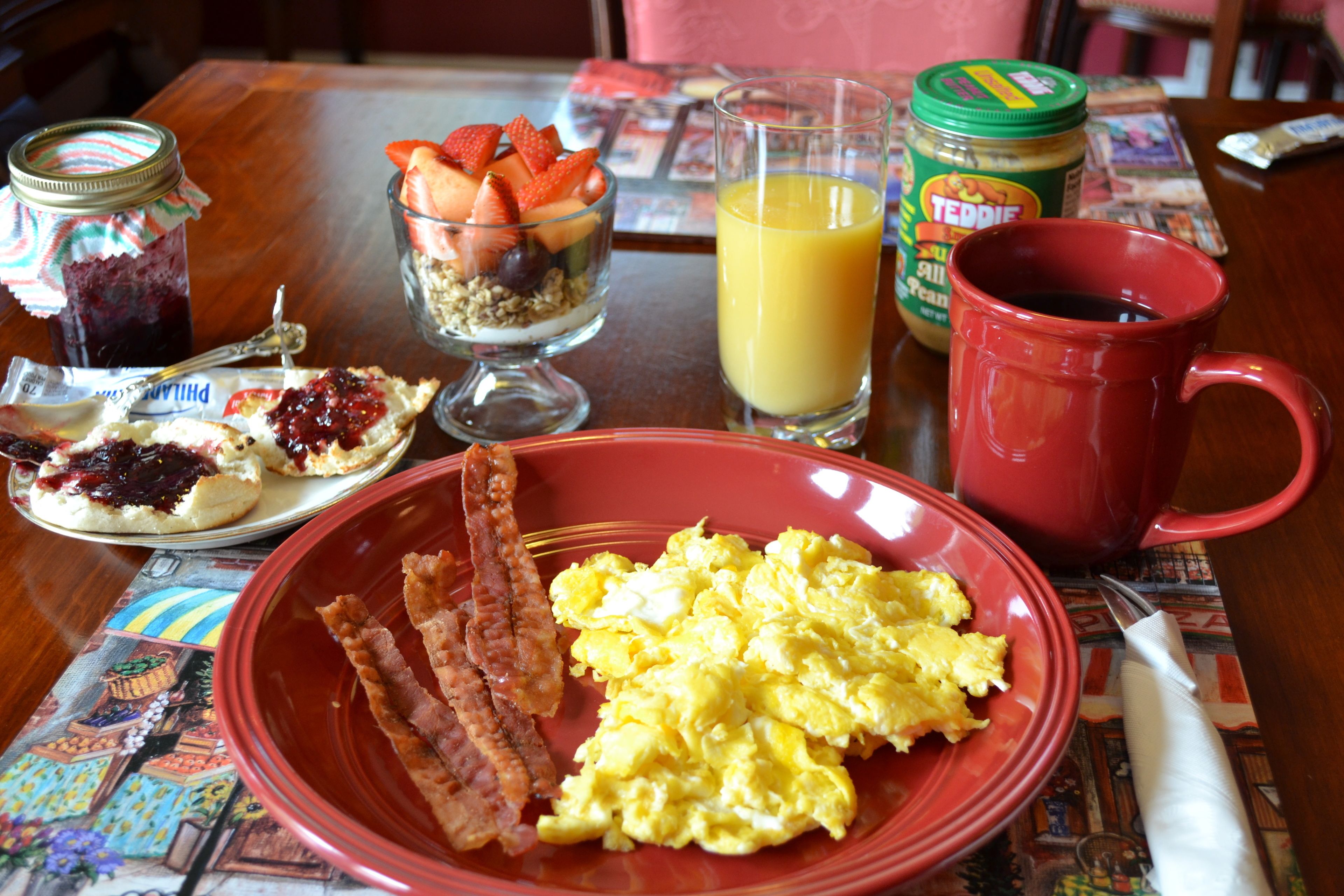 Una tavola imbandita per la colazione con uova strapazzate, pancetta, un bicchiere di succo d'arancia, un vasetto di burro di arachidi e una tazza rossa.