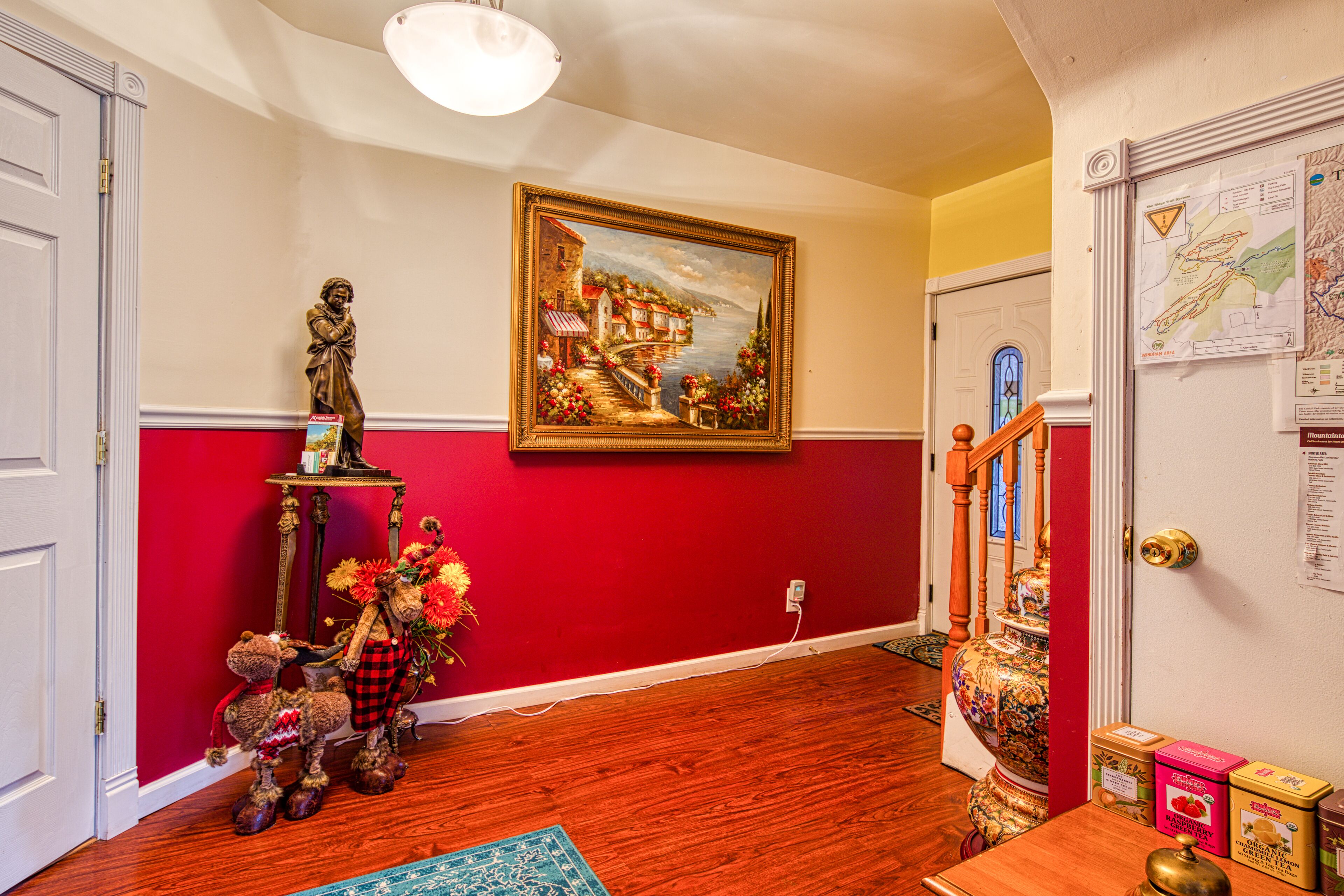 A hallway with a red wall, a wooden floor, a painting, a statue, a table with a vase of flowers, and a door with a map on the wall.