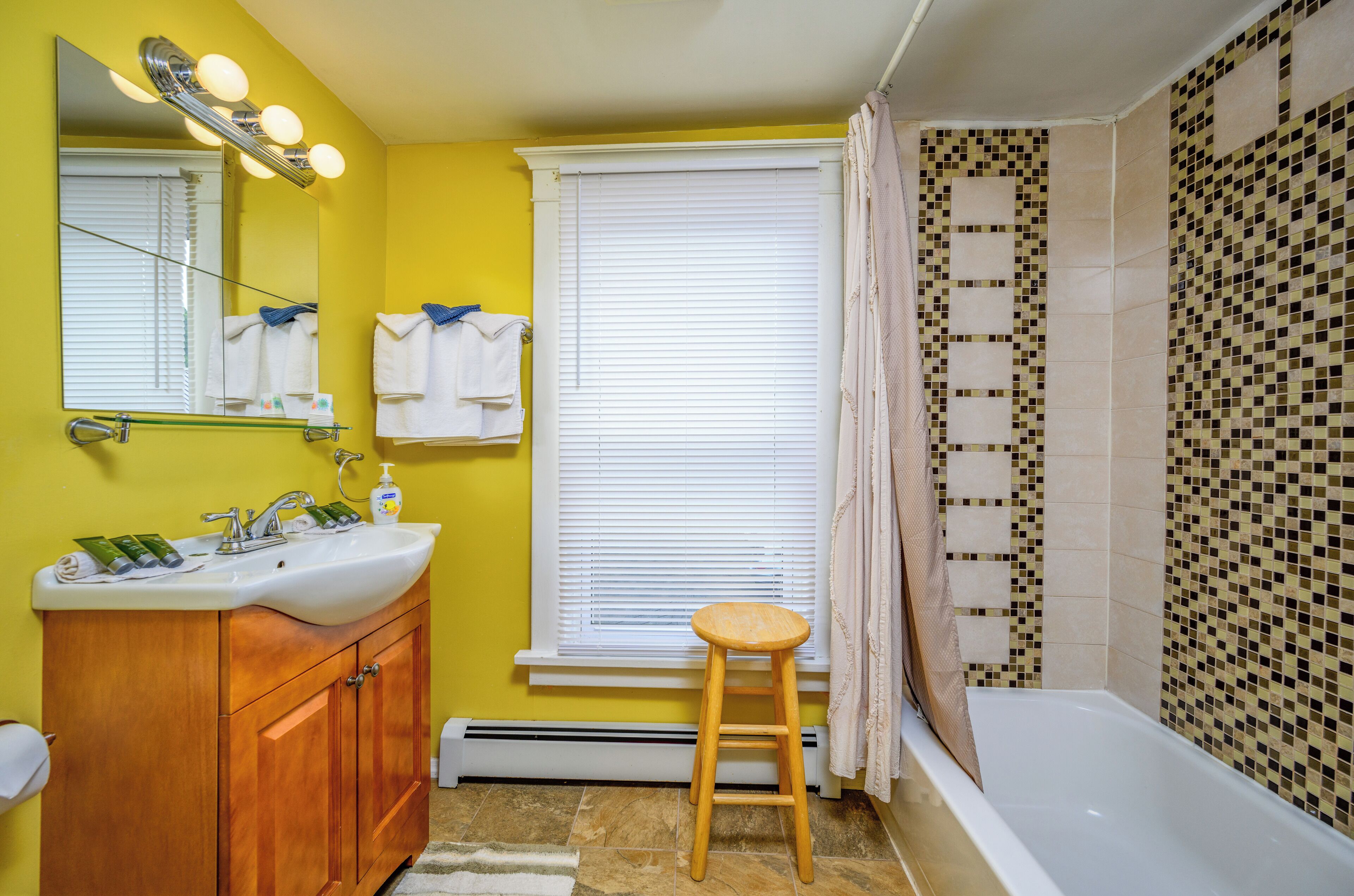 A bathroom with yellow walls, a wooden vanity with a sink, a mirror with lights, a window with blinds, a shower with mosaic tiles, and a wooden stool.