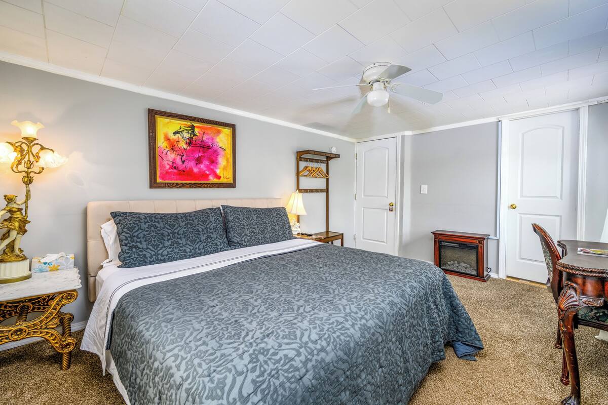 A bedroom with a large bed, a ceiling fan, a painting, a lamp, and a small table with a statue.