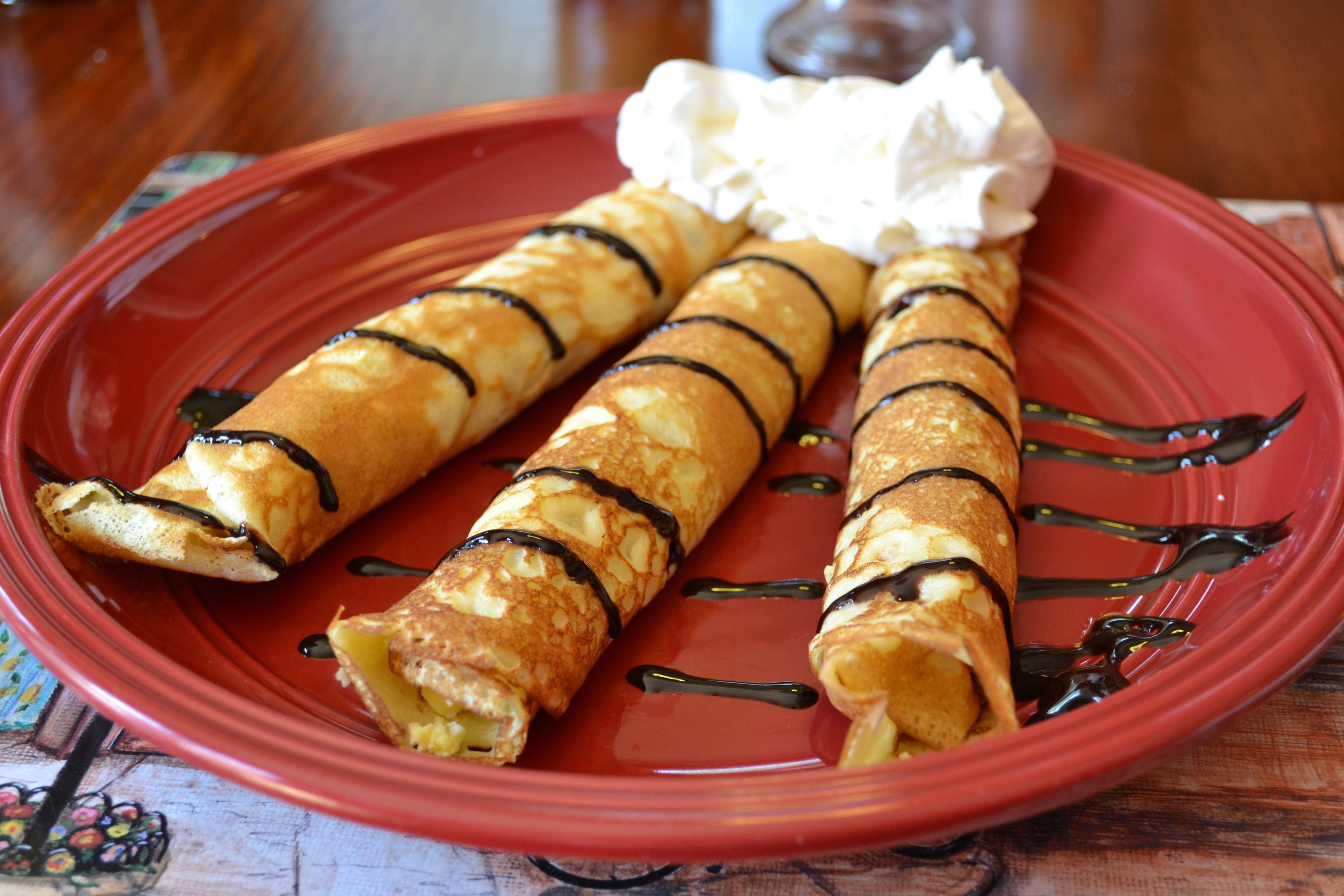 A red plate with three crepes topped with chocolate sauce and whipped cream.