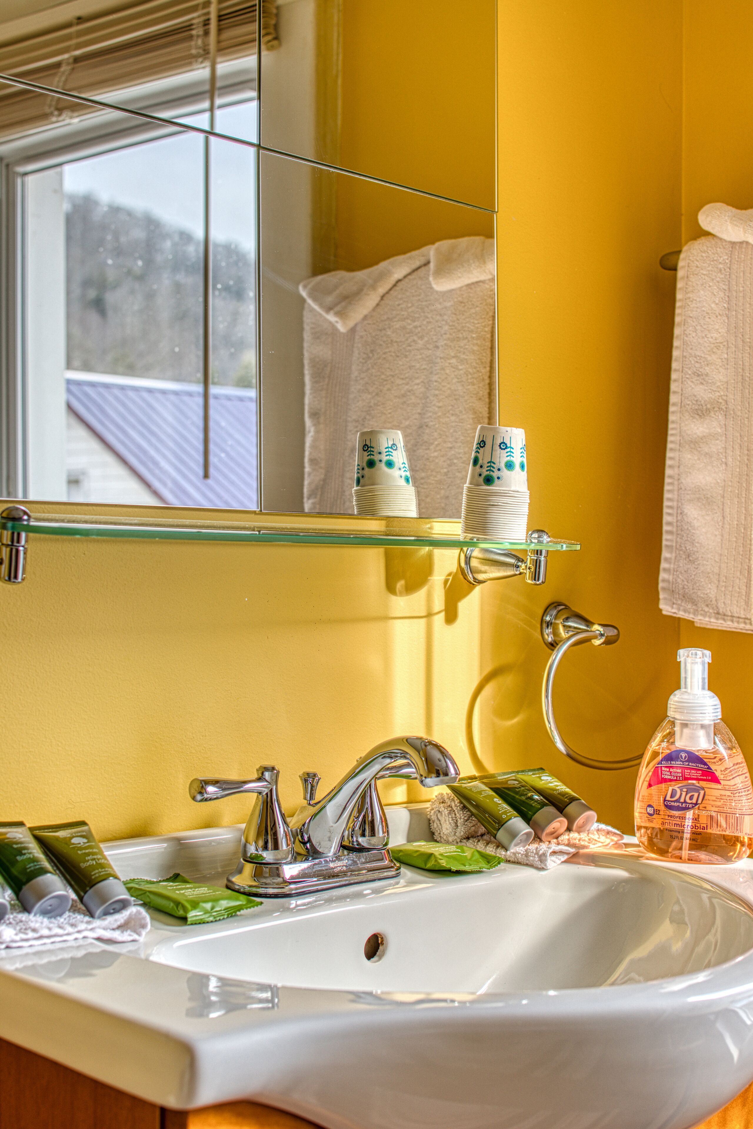 Une salle de bain avec un lavabo blanc, des murs jaunes et une étagère en verre au-dessus du lavabo où sont rangés les articles de toilette.