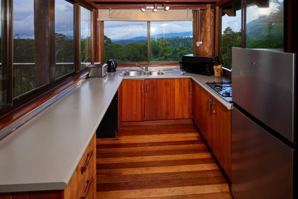 A modern kitchen with wooden cabinets, a stainless steel refrigerator, and a large window overlooking a forested landscape.