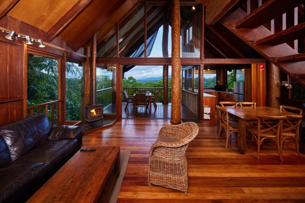 A spacious room with a wooden ceiling, a fireplace, a dining area with a table and chairs, and a large window offering a view of greenery.