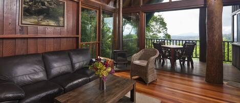 A wooden interior with a leather sofa, a wooden coffee table with a vase, wicker chairs, and a view through large windows.