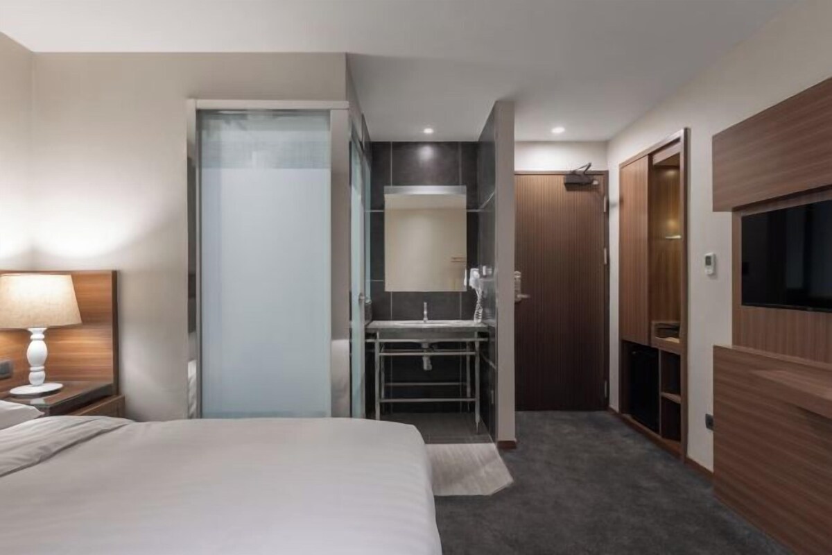 Een moderne hotelkamer met een bed, een nachtkastje met een lamp, een badkamer met een spiegel en een aan de muur bevestigde televisie.