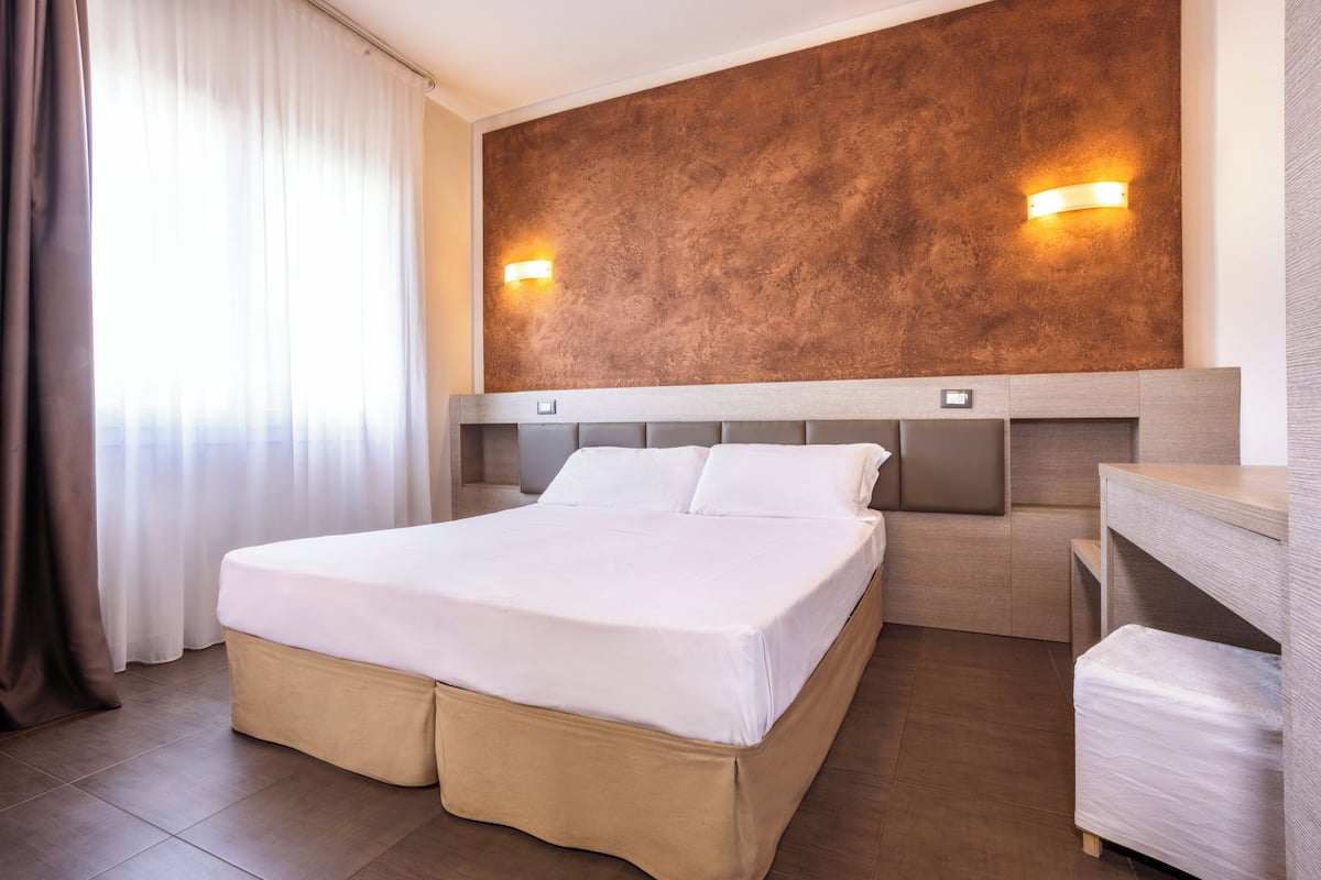 A hotel room with a large bed, a wooden headboard, two wall-mounted lights, a bedside table, and a window with curtains.