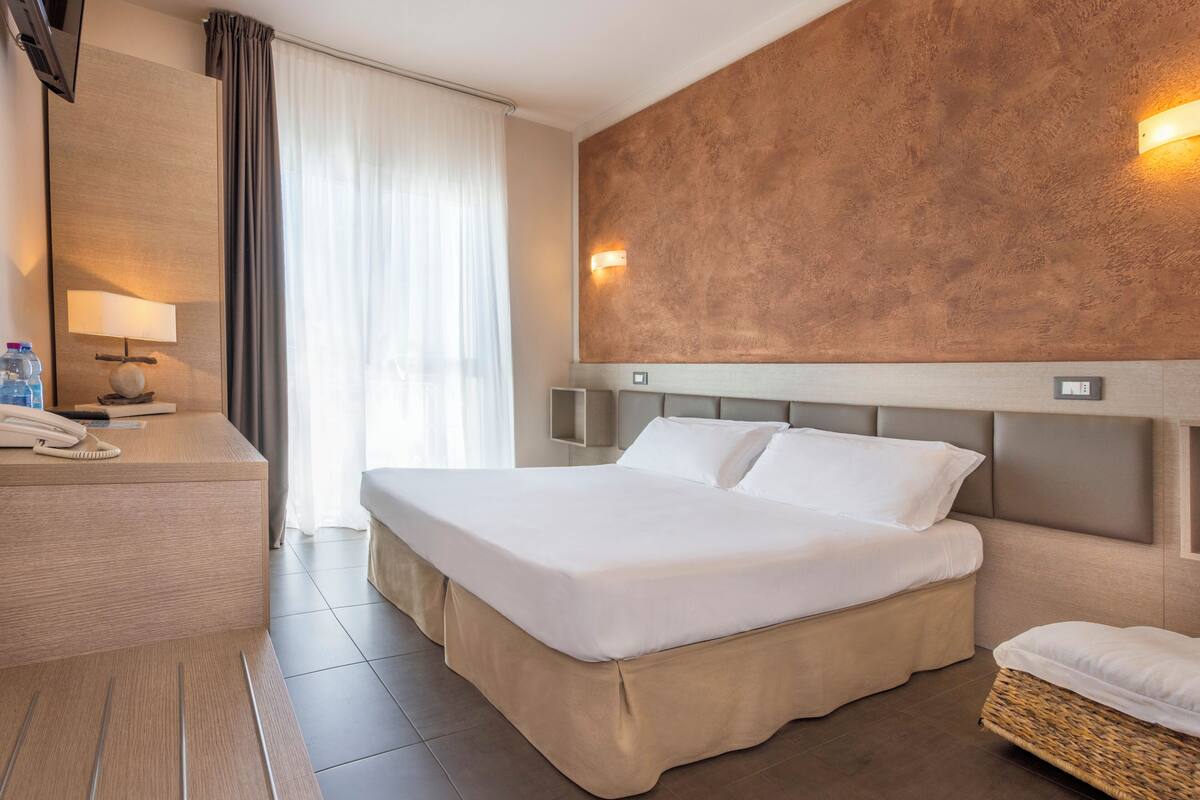 A hotel room with a large bed, a bedside table with a lamp, a wall-mounted TV, and a large window with curtains.