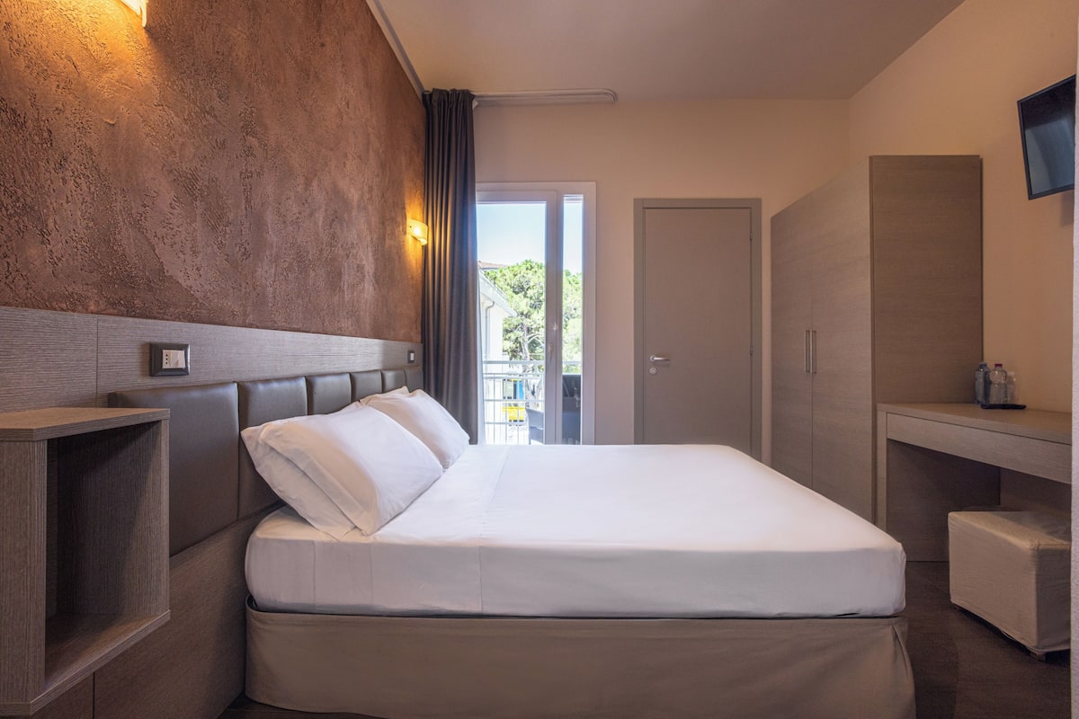 A hotel room with a bed, bedside table, wardrobe, and a view of the outdoors.