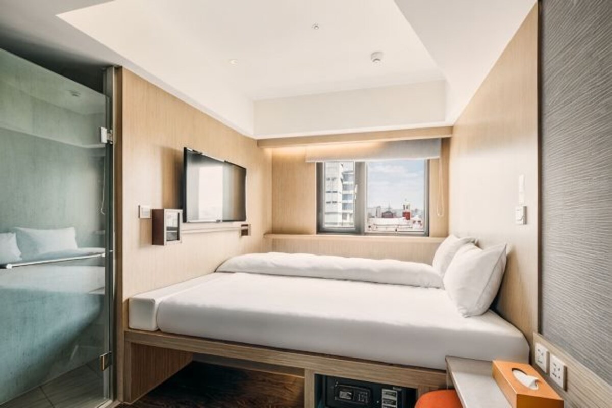 A modern hotel room with a bed, a TV, a window with a city view, and a shower area.