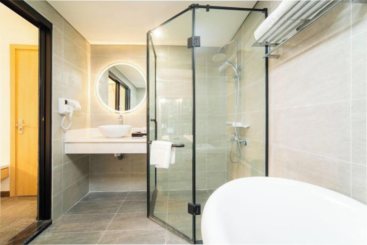 A modern bathroom with a glass-enclosed shower, a white sink, a round mirror, and a white bathtub.