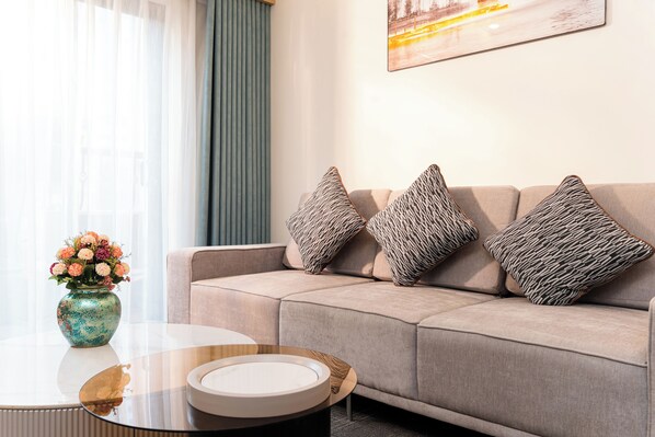 A modern living room with a beige sofa, patterned cushions, a glass coffee table, and a vase of flowers. - Capital Garden Hotel (Hanoi)