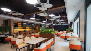 A modern dining area with hexagonal ceiling lights, orange and white chairs, and a bar area in the background. - Capital Garden Hotel (Hanoi)