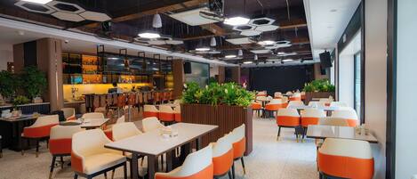A modern dining area with hexagonal ceiling lights, orange and white chairs, and a bar area in the background.
