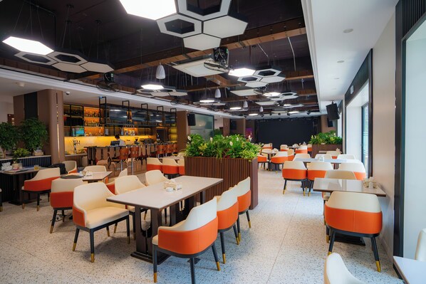 A modern dining area with hexagonal ceiling lights, orange and white chairs, and a bar area in the background. - Capital Garden Hotel (Hanoi)