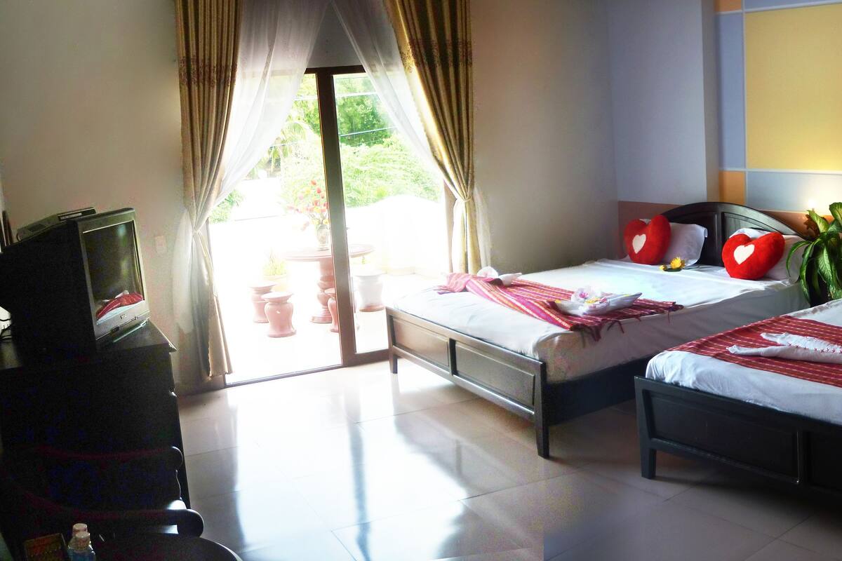 Ein Hotelzimmer mit zwei Betten, jedes mit roten herzförmigen Kissen und weißen Handtüchern. Links befindet sich ein Fernseher auf einem Tisch, rechts ein Fenster mit Vorhängen.