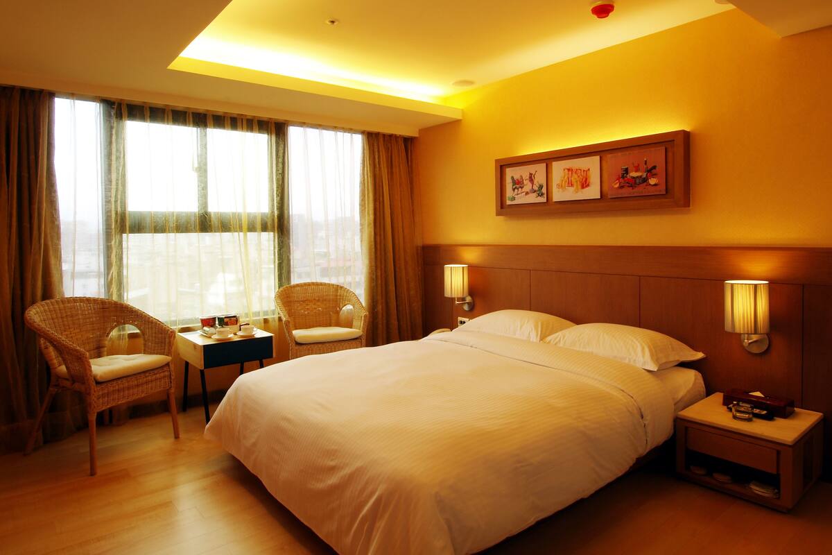 Habitación de hotel con una cama grande, dos mesitas de noche, una silla y una ventana con cortinas.