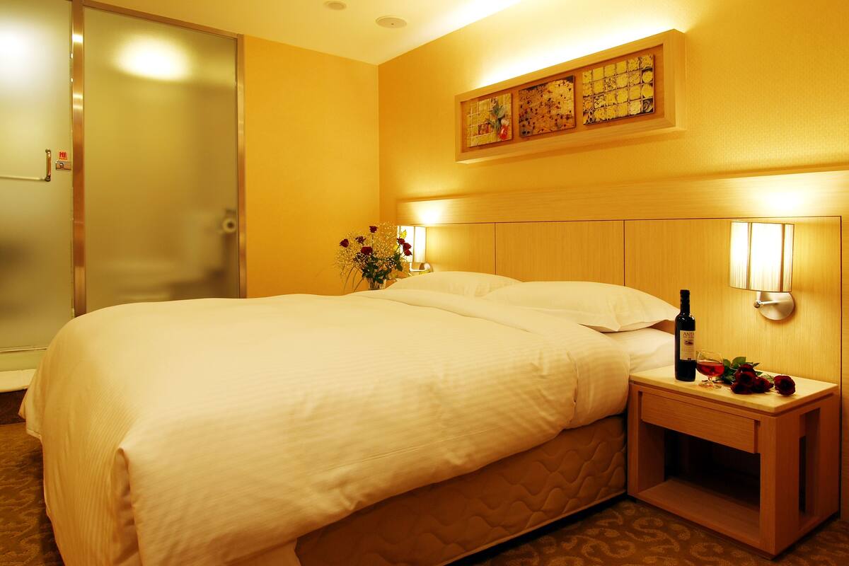 Habitación de hotel con una cama grande, una mesita de noche con una botella de vino y copas, un jarrón con flores y un cuadro en la pared.