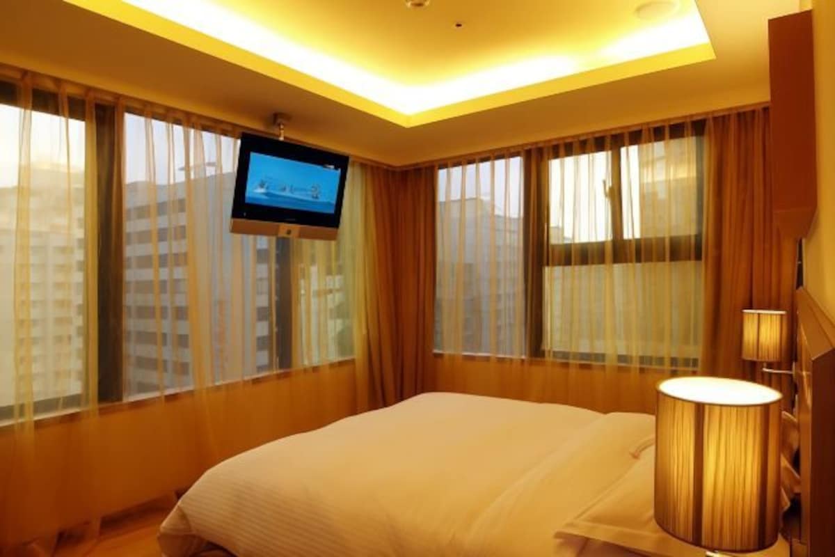 Habitación de hotel con cama, televisor montado en la pared y un ventanal con cortinas.