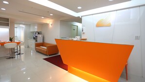 A modern reception area with a bright orange desk, white walls, and a visible kitchen area in the background. - Morwing Hotel Fuzhong (New Taipei City)