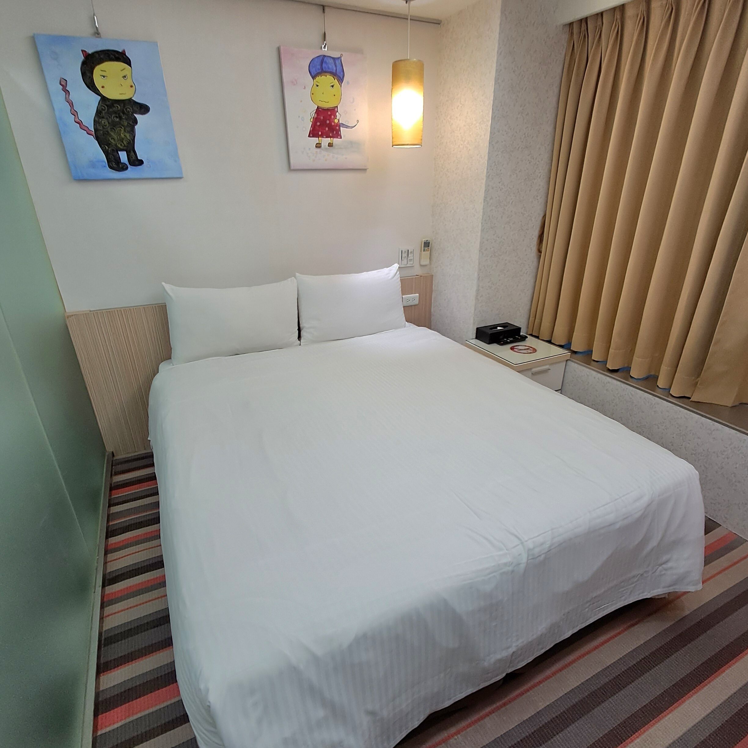 A hotel room with a large bed, two pillows, a bedside table with a phone and a lamp, a striped rug, and two wall art pieces.