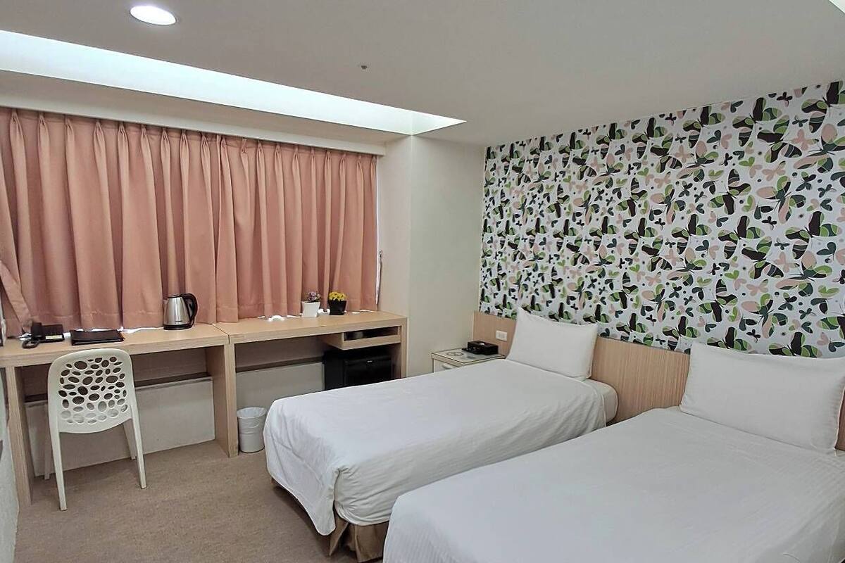 Une chambre d’hôtel avec deux lits, un bureau, une chaise, une fenêtre avec des rideaux et un mur décoré d’un motif.