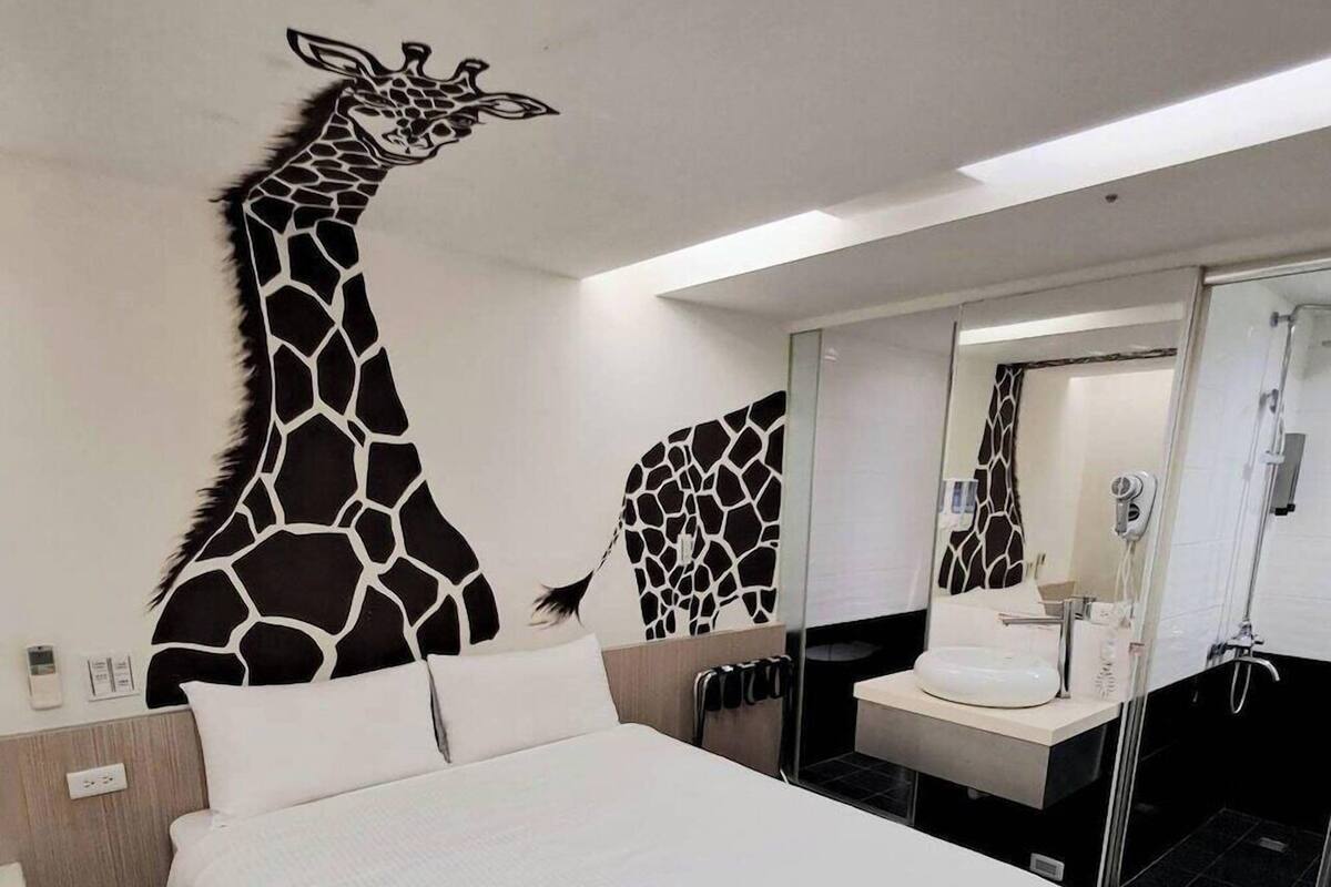 Une chambre d’hôtel avec un grand lit, une salle de bain moderne dotée d’une douche en verre et une décoration murale sur le thème des girafes.