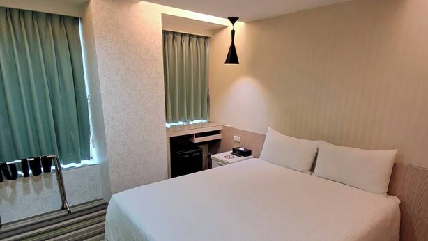 A hotel room with a large bed, a bedside table, a lamp, a window with curtains, and a striped carpet. - Morwing Hotel Fuzhong (New Taipei City)
